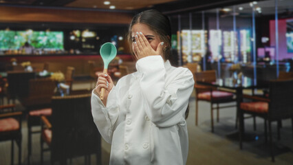 Woman chef holding teal spoon and covering face with hand while smiling in a restaurant building;...