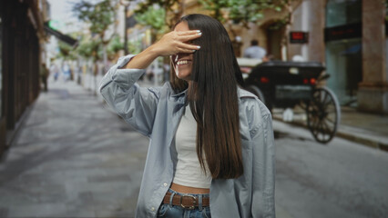 Fototapeta premium Young woman covering eyes with hand and smiling on a narrow street near a carriage; playful surprise.
