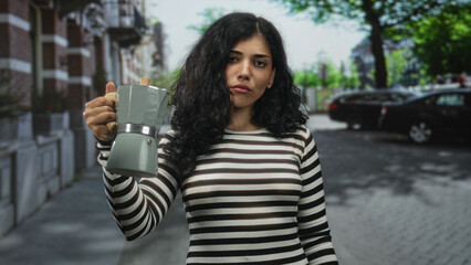 Fototapeta premium Young arab woman holding moka pot in outstretched bare hand on a cobbled street by brick building, striped top visible; quiet contemplation.