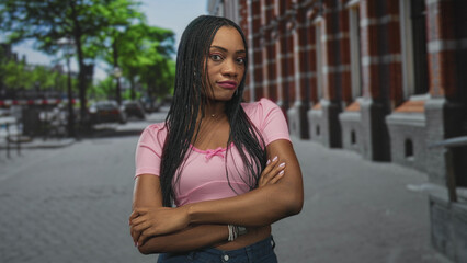 Fototapeta premium Woman with arms crossed, braided hair, pink cropped top and jeans, bracelets and nails visible, standing on a street beside a brick building and parked cars; disapproval.