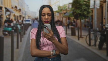 Woman in pink top holds smartphone and scrolls with one hand while arms are crossed, looking at the screen on a busy street; pensive reflection.