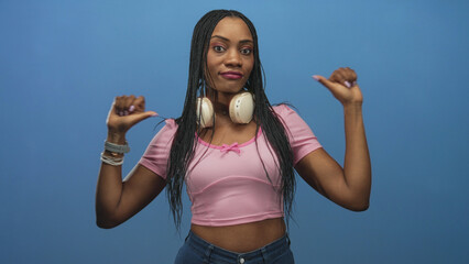 Black woman in pink crop top with headphones around neck points thumbs to chest against a studio blue backdrop, braided hair and bracelets visible; self confidence empowerment.