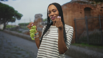 Woman holding a bunch of green grapes, showing fingers gesture on a cobblestone street beside...