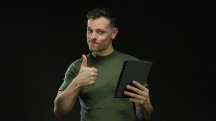 Young man interacting with tablet while smiling on black background showing approval with thumb up...
