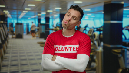 Young man wearing volunteer shirt making funny faces in gym with exercise equipment in background, showcasing humorous expression in an active setting.