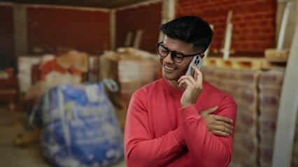 Man in red sweater and glasses smiling while talking on smartphone in indoor construction site with blurred materials around, suggesting a busy and productive environment.