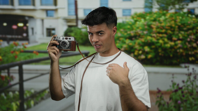 Young man holding a vintage camera outdoors in a city environment surrounded by greenery showcasing a relaxed and confident pose under natural sunlight. - Powered by Adobe
