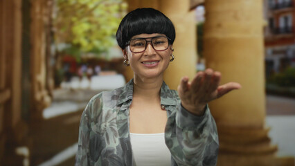 Woman smiling outdoors offering hand wearing glasses and blouse standing near columns in sunny...