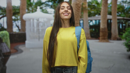Fototapeta premium Teenage girl in yellow sweater with blue backpack standing with head tilt and subtle smile in park; youthful optimism.
