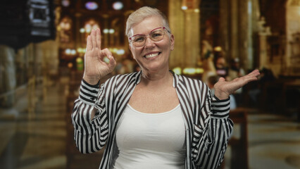Middle aged woman smiling, showing ok sign with raised hands inside a church building interior,...