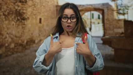 Woman with glasses and red backpack thumbs pointing to chest on old town street by stone archway; surprise self identity.