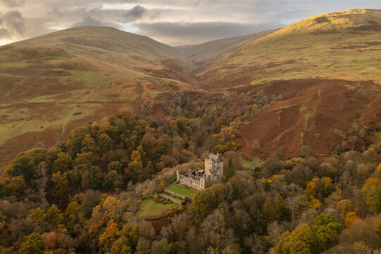 Aerial view of Castle Campbell nestled in a vibrant autumn forest, contrasting with the rolling ochre hills under a cloudy sky, Dollar, Scotland, United Kingdom.