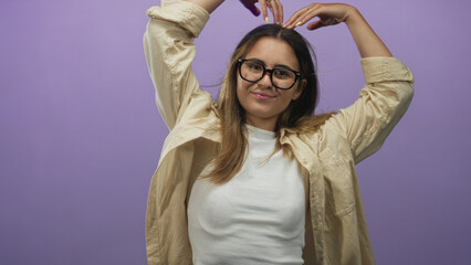 Hispanic woman wearing glasses and beige overshirt over white tee making heart gesture with hands above head in purple studio; playful.