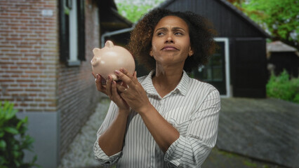 Woman smiling holding piggybank, finger to chin thinking at house entrance; savings planning...