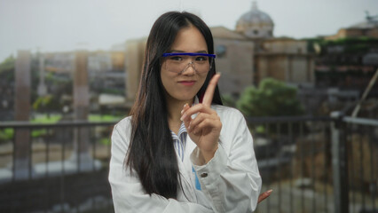 Woman scientist in lab coat with glasses gestures outdoors against ancient roman ruins background using finger and hand to express refusal or denial.