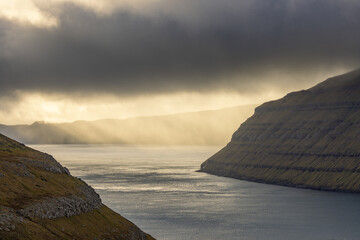 View of sun rays pierce through dramatic clouds, illuminating the tranquil waters nestled between rugged, textured cliffs, Sorvagur, Vagar, Faroe Islands.