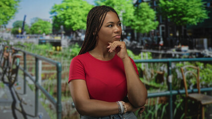 Fototapeta premium Woman with hand on chin on street canal railing in green park wearing red tshirt and bracelet; thoughtful contemplation.