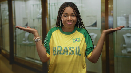 Woman shrugging with palms up in jewelry showcase building, wearing brasil shirt and bracelets; uncertainty doubt.