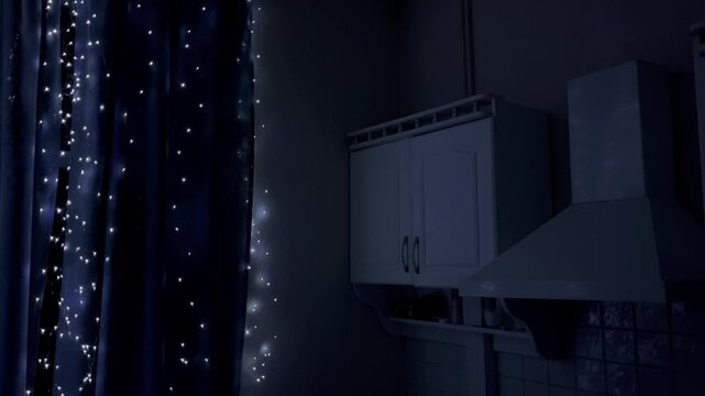 Dimly lit cozy kitchen with white cabinets, range hood, tiled backsplash, and blue glow from string fairy lights near dark curtains. High quality 4k footage