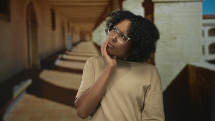 Fototapeta premium Woman in beige shirt and clear glasses rests chin on hand under arched corridor of old stone building; contemplation reflection introspection doubt.