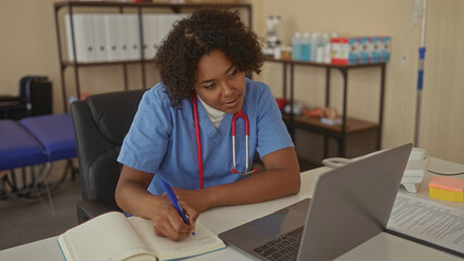 Woman writing notes in notebook with blue pen at clinic desk beside an open laptop; quiet...