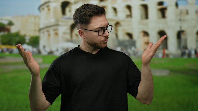 Young man with beard looking at his hands in front of colosseum in rome, appearing thoughtful and indecisive in the iconic outdoor roman setting, italy.