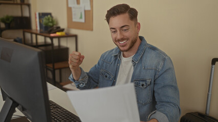 Young man with beard working in office, focused on computer screen, wearing denim jacket and reading papers in a professional indoor setting with shelves in background.