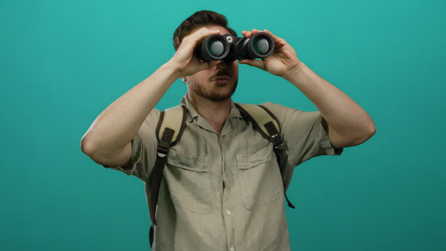 Young man with beard wearing backpack and looking through binoculars against isolated green background in casual tourist exploration scene.