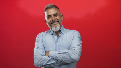 Man smiling with arms crossed wearing a light blue shirt and grey beard in a red studio; confidence warmth approachability.