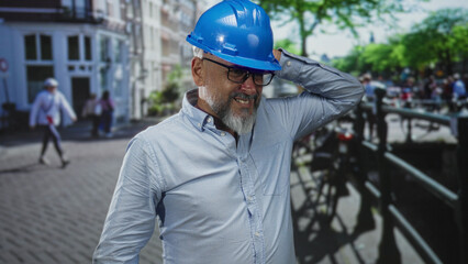 Man architect in blue hard hat and glasses adjusting helmet with hand while standing on a city street; steady professional confidence.