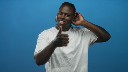 Man in white t shirt holding hand to ear and giving thumbs up in studio with solid blue backdrop; playful confidence.