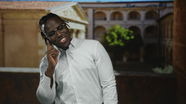 Young african american man holding smartphone to ear, smiling and gesturing on an old town street; joy. - Powered by Adobe