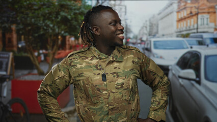 Man in camouflage soldier uniform with dogtags, young and smiling with hands on hips on a busy city street with parked cars; pride duty camaraderie.