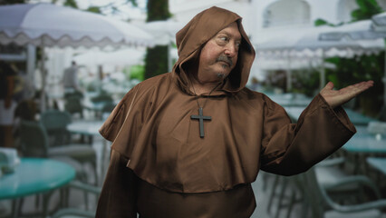 Naklejka premium Man in brown robe with cross pendant, palm up gesture at an outdoor cafe terrace, presenting a welcome pose; serenity.