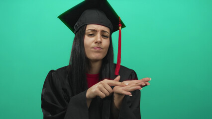 Hispanic woman in graduation gown gestures for money against green wall, expressing financial need after graduation in isolated, academic setting.