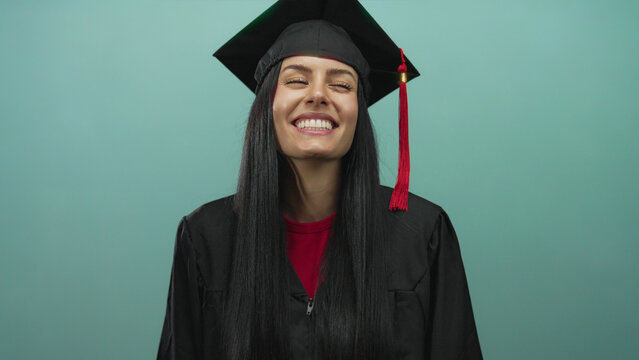 Young hispanic woman in graduation gown and cap smiles joyfully against a blue wall, symbolizing academic achievement and success.