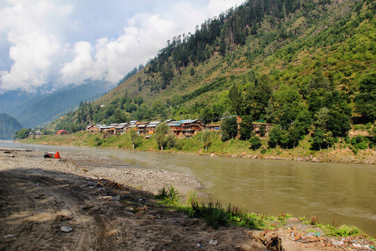 View of a serene river reflecting the sky, with wooden houses nestled at the base of lush, green mountains, a tranquil scene in Neelum Valley, Azad Kashmir, Pakistan.