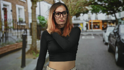Young woman touching her shoulder and showing bare midriff, wearing oversized glasses on a street; pensive solitude.