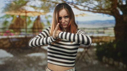 Young redhead woman pressing hands together in a defensive stop gesture on a sunlit terrace at a building; defiance.