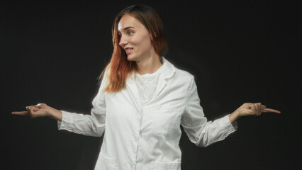 Young woman wearing a white lab coat points both index fingers outward while smiling toward camera...