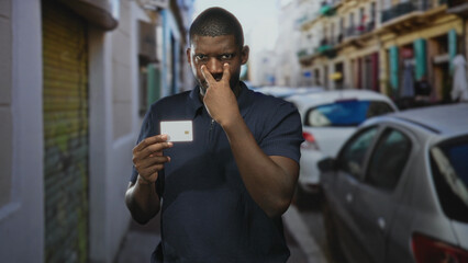 Young black man holds creditcard and points finger at camera in narrow street with parked cars and...