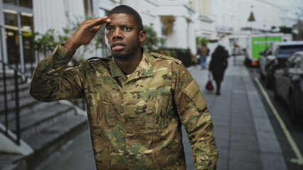 Fototapeta premium Man soldier in camouflage uniform with hand to forehead shading eyes on a city street beside parked cars and building steps, looking ahead; duty vigilance.