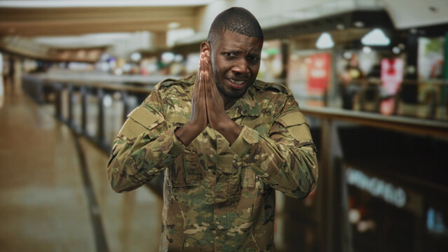 Soldier man in camouflage uniform pressing hands together near storefronts on airport concourse in pleading gesture; plea anxiety.