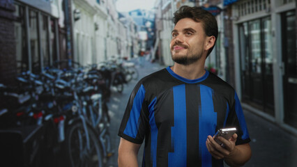 Man holding smartphone on amsterdam street, smiling and looking up while wearing a blue and black jersey; optimism exploration.