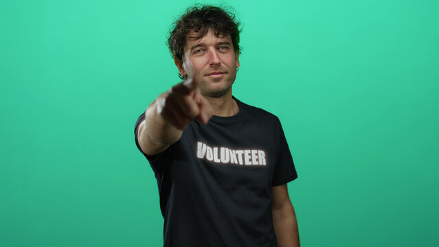 Hispanic man in volunteer shirt pointing forward against green background, conveying enthusiasm and determination for service and helping others with a confident gesture.