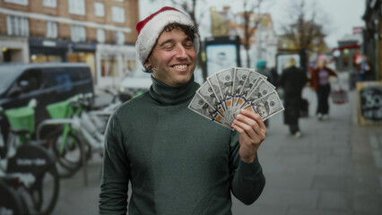 Fototapeta premium Man wearing christmas hat smiling while holding dollars on street outdoors during holiday season with blurred background and people walking by.
