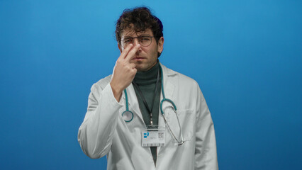 Hispanic doctor with glasses pointing and making vigilant gesture against blue background, dressed...