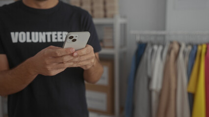 Hispanic volunteer man in black shirt using smartphone in organized room with shelves and clothes, symbolizing tech-savvy aid work in a structured environment.