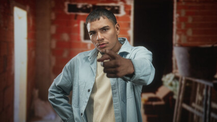 Man points finger at camera inside a raw brick building with ladder, scaffolding and loose bricks, young hispanic male in light denim shirt; challenge defiance.