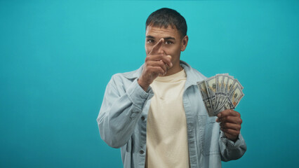 Young hispanic man holding fanned us dollars and pointing with fingers at camera in studio;...